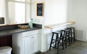 Een keuken met een bar, witte kastjes, een welkomstbord en barstoelen.