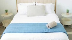 A neatly made bed with a wooden headboard, white bedding, and a blue blanket. There are two bedside tables with small potted plants.