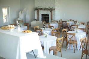 Un espace repas avec des tables rondes et des chaises en bois, une nappe blanche et une cheminée agrémentée de plantes décoratives.