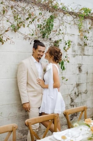 A couple standing against a wall with climbing plants, wooden chairs, and a table with a floral arrangement. - -The Gathered Place- (Groveland)