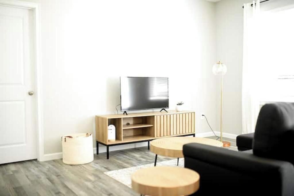 Ett modernt vardagsrum med en platt-TV på ett träställ, två runda träbord, en svart soffa och en golvlampa.