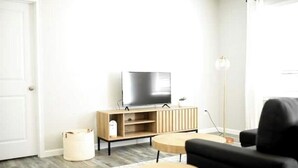 A modern living room with a flat-screen TV on a wooden stand, two round wooden tables, a black sofa, and a floor lamp.