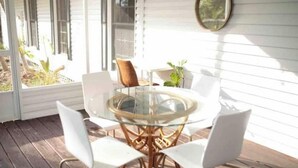 Een ronde glazen tafel met witte stoelen op een houten terras, omringd door witte muren en een raam met zonwering.