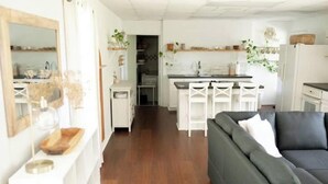 Een moderne woonkamer met een donkere bank, een houten bijzettafeltje en een witte salontafel. In de achtergrond is een keuken met witte kasten en een eethoek met witte stoelen te zien.