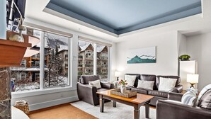 A modern living room with a sofa, coffee table, and a large window with a view of buildings and trees. - Exciting Rockies 1BR Escape, Hot Tub, Pool & BBQ! (Canmore)