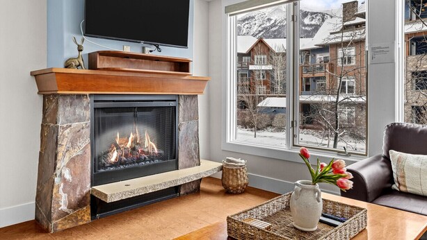 A fireplace with a stone surround, a flat-screen TV mounted above it, and a view of a snowy mountain landscape through a large window. - Exciting Rockies 1BR Escape, Hot Tub, Pool & BBQ! (Canmore)