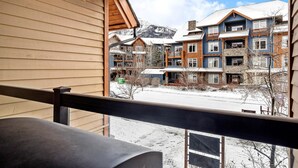 A view from a balcony with a wooden railing, overlooking a multi-story building with a snow-covered landscape and mountains in the background. - Exciting Rockies 1BR Escape, Hot Tub, Pool & BBQ! (Canmore)