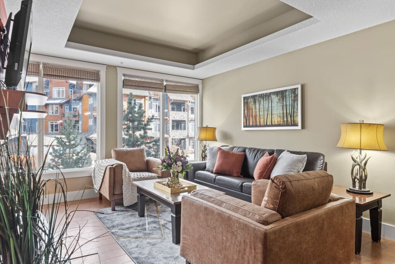A living room with a sofa, armchairs, a coffee table, and a TV. There are two lamps, a painting on the wall, and a window with a view of buildings and trees.