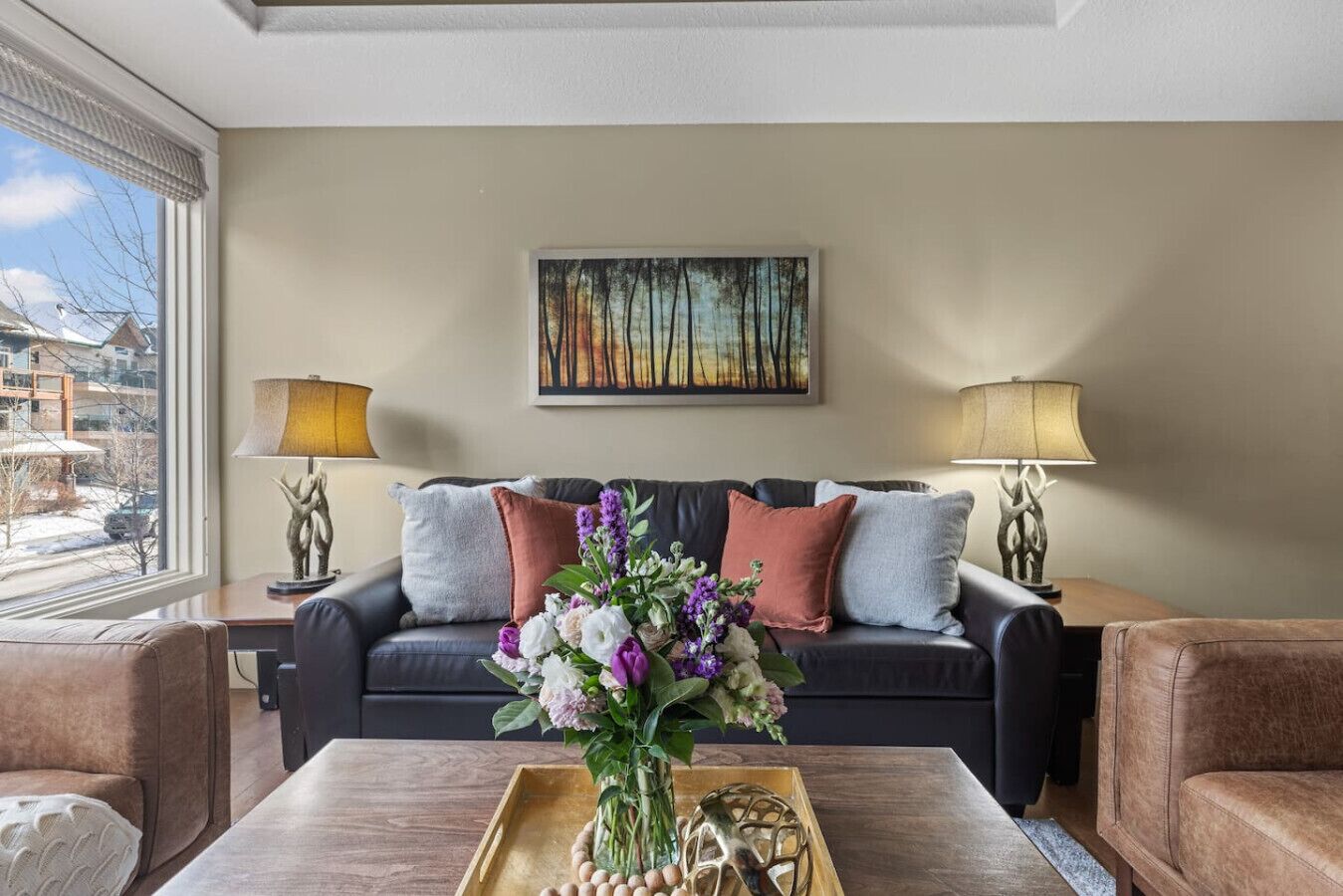A modern living room with a black leather sofa, two side tables with lamps, a wooden coffee table with a vase of flowers, and a painting above the sofa.