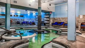 Una piscina interior con múltiples jacuzzis, zonas de estar azules y una fuente central.