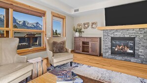 A cozy living room with a fireplace, a leather sofa, a wooden coffee table, and a view of mountains through large windows. - Serenity Awaits | Mountain Views (Canmore)