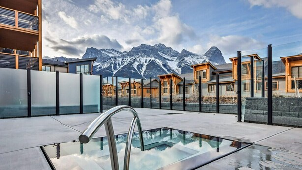 A rooftop pool with a glass barrier, modern buildings, and a mountain backdrop. - Serenity Awaits | Mountain Views (Canmore)