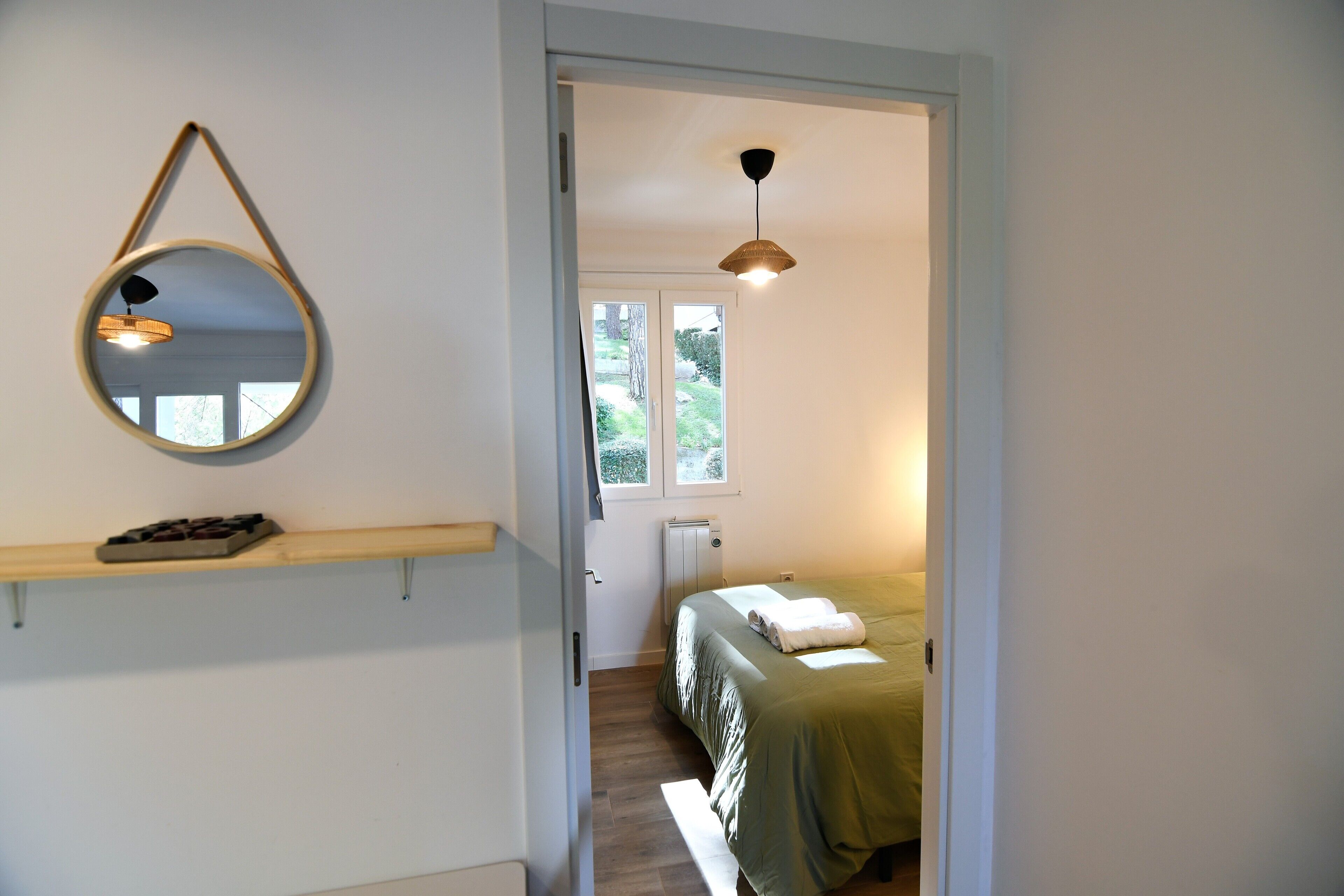 Un dormitorio con cama, un espejo colgado, una estantería con bombones y una ventana con luz natural.
