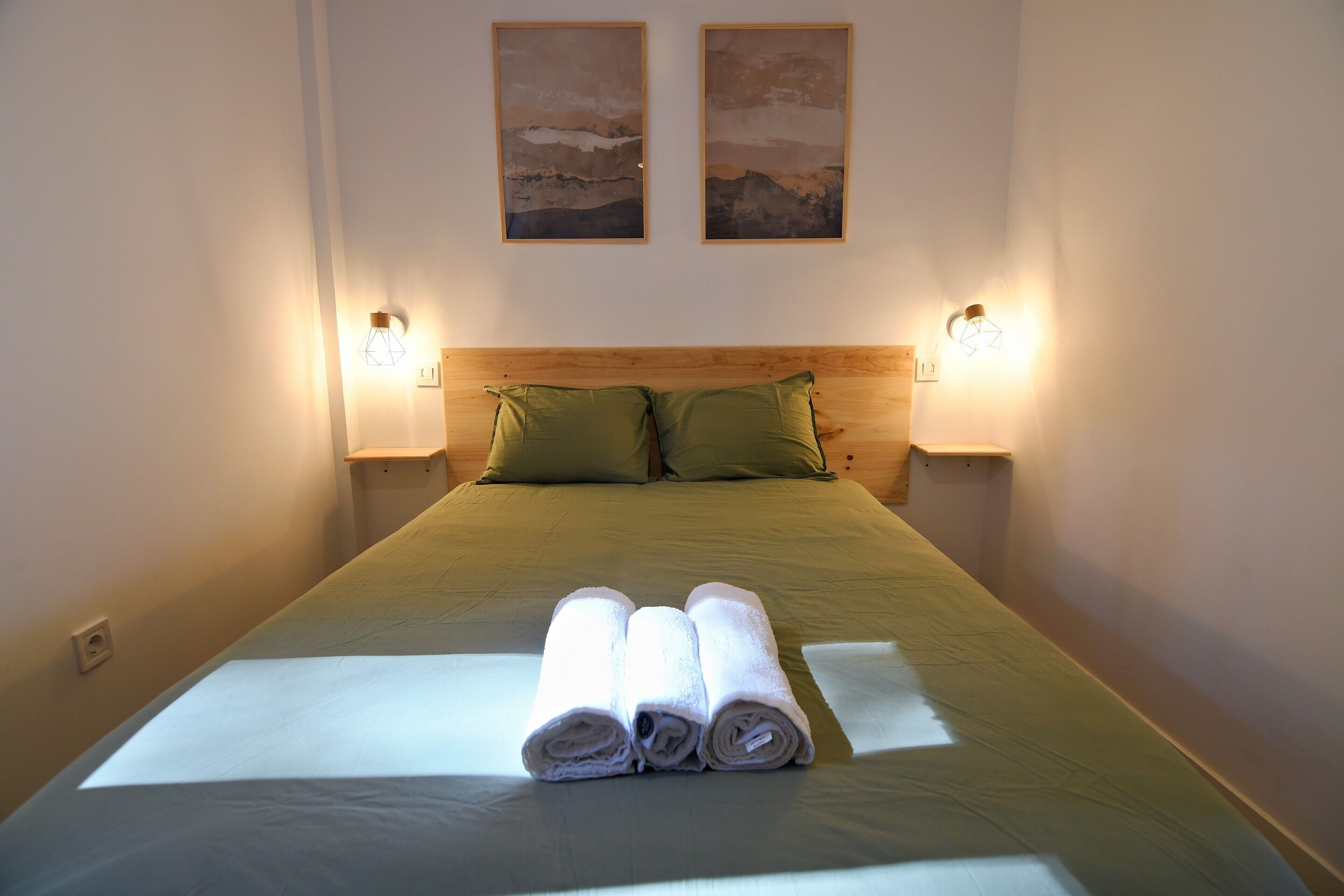 A neatly made bed with green bedding and two pillows, a bedside table with a lamp, and two framed pictures on the wall.