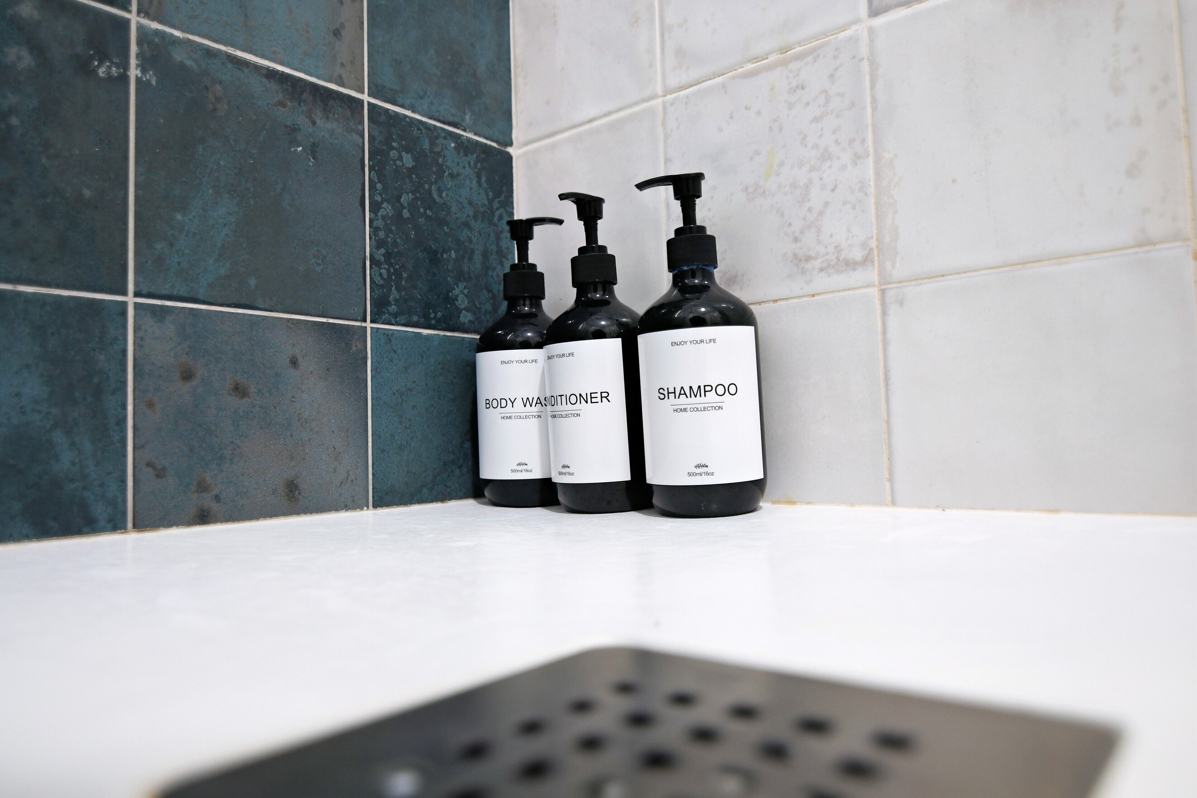 Tres botellas negras de gel de baño y champú sobre una superficie blanca, frente a una pared de azulejos.