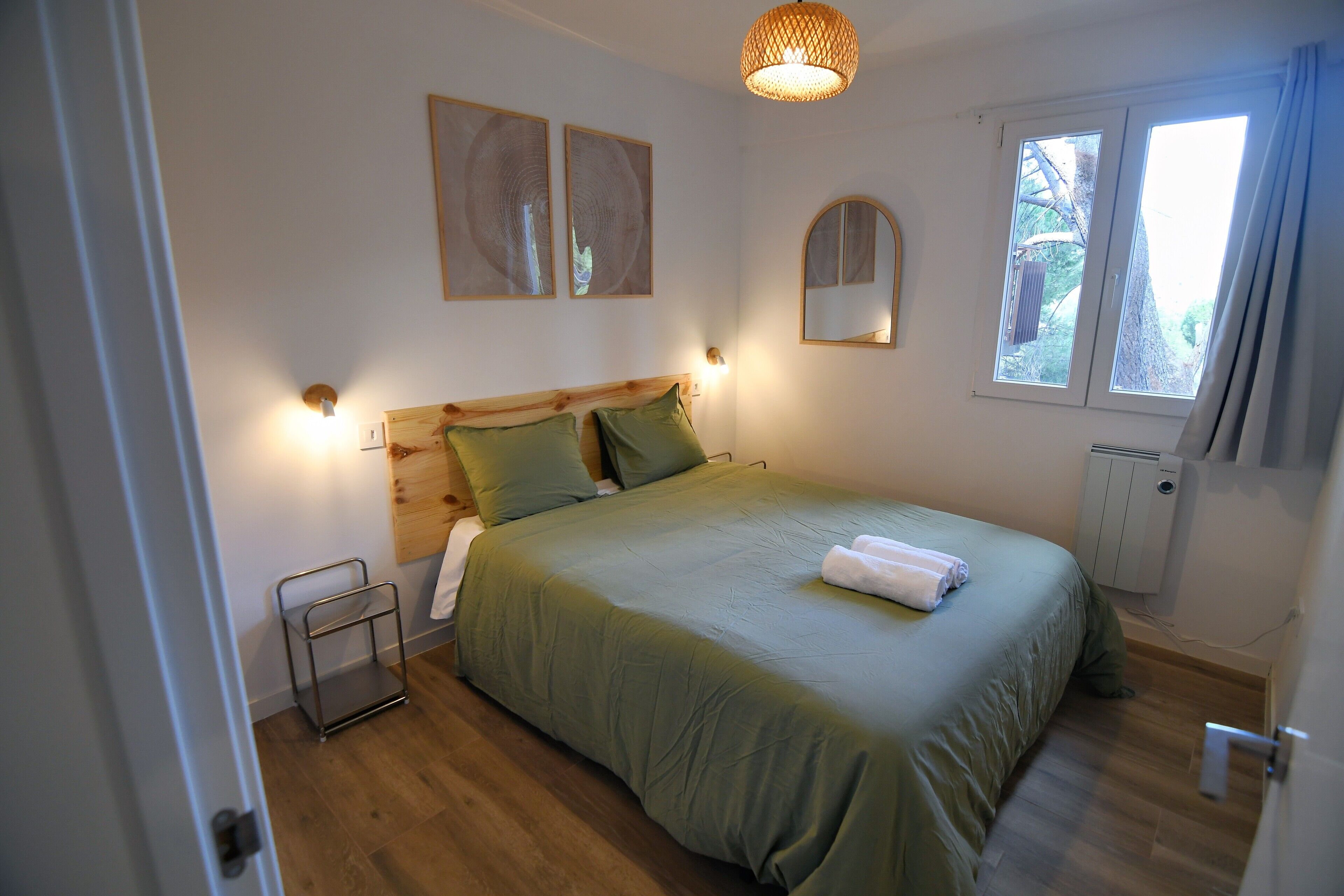 A bedroom with a wooden headboard, green bedding, a bedside table, a wall-mounted mirror, and a window with curtains.