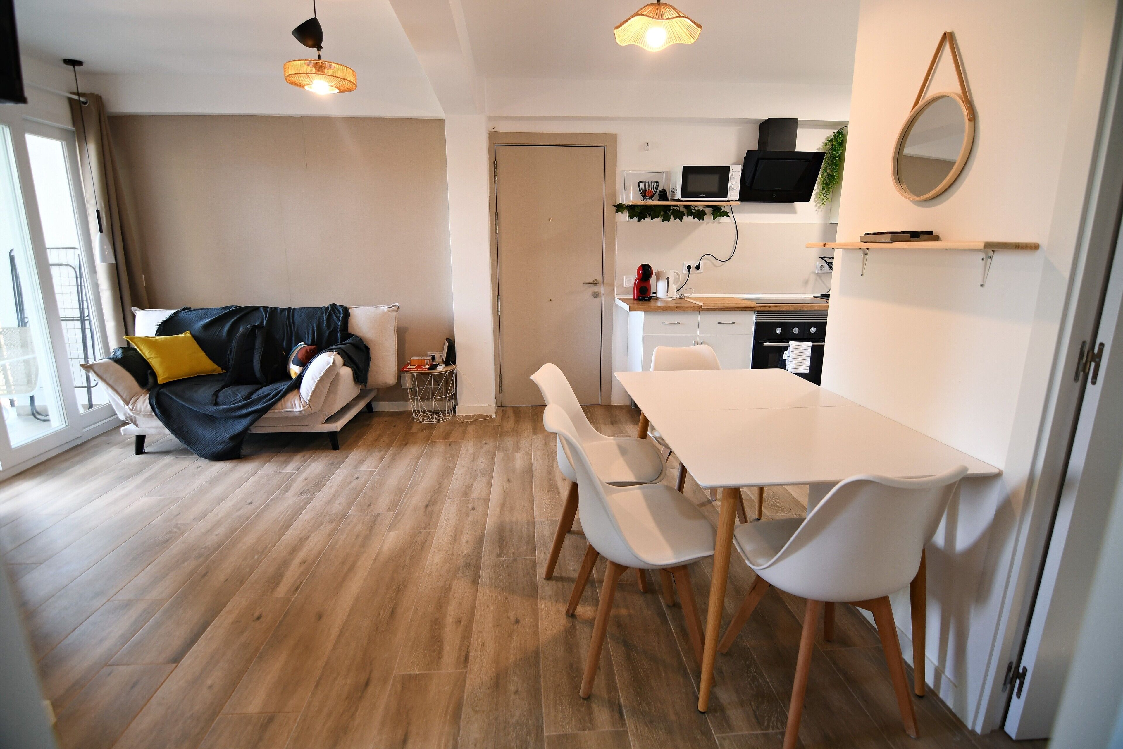 Una sala moderna con sofá, mesa de comedor y sillas. Cuenta con una zona de cocina con microondas y fregadero. El piso es de madera y hay un ventanal grande.