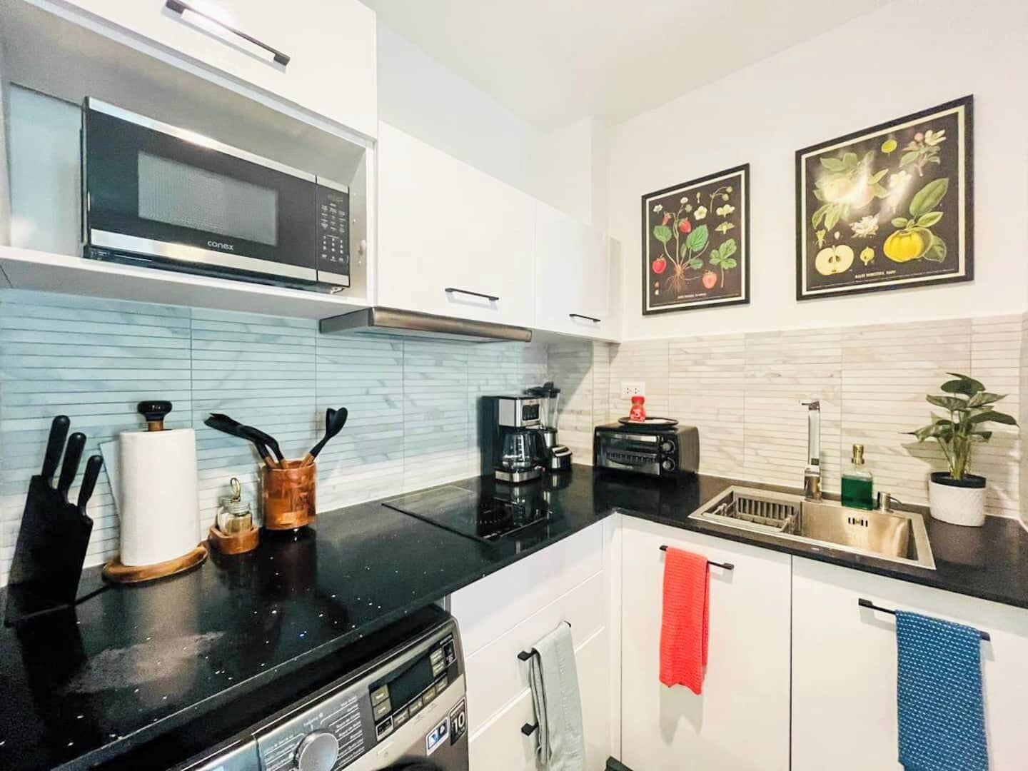 Eine moderne Küche mit weißen Schränken, einer schwarzen Arbeitsplatte, einer Mikrowelle, einer Kaffeemaschine und zwei gerahmten Pflanzenbildern an der Wand.