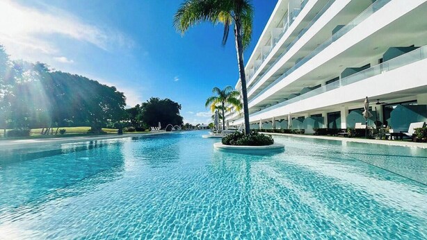 A clear swimming pool with a palm tree in the center, surrounded by a modern building with multiple balconies. - Penthouse Exclusivo Cerca del Hard Rock (Punta Cana)