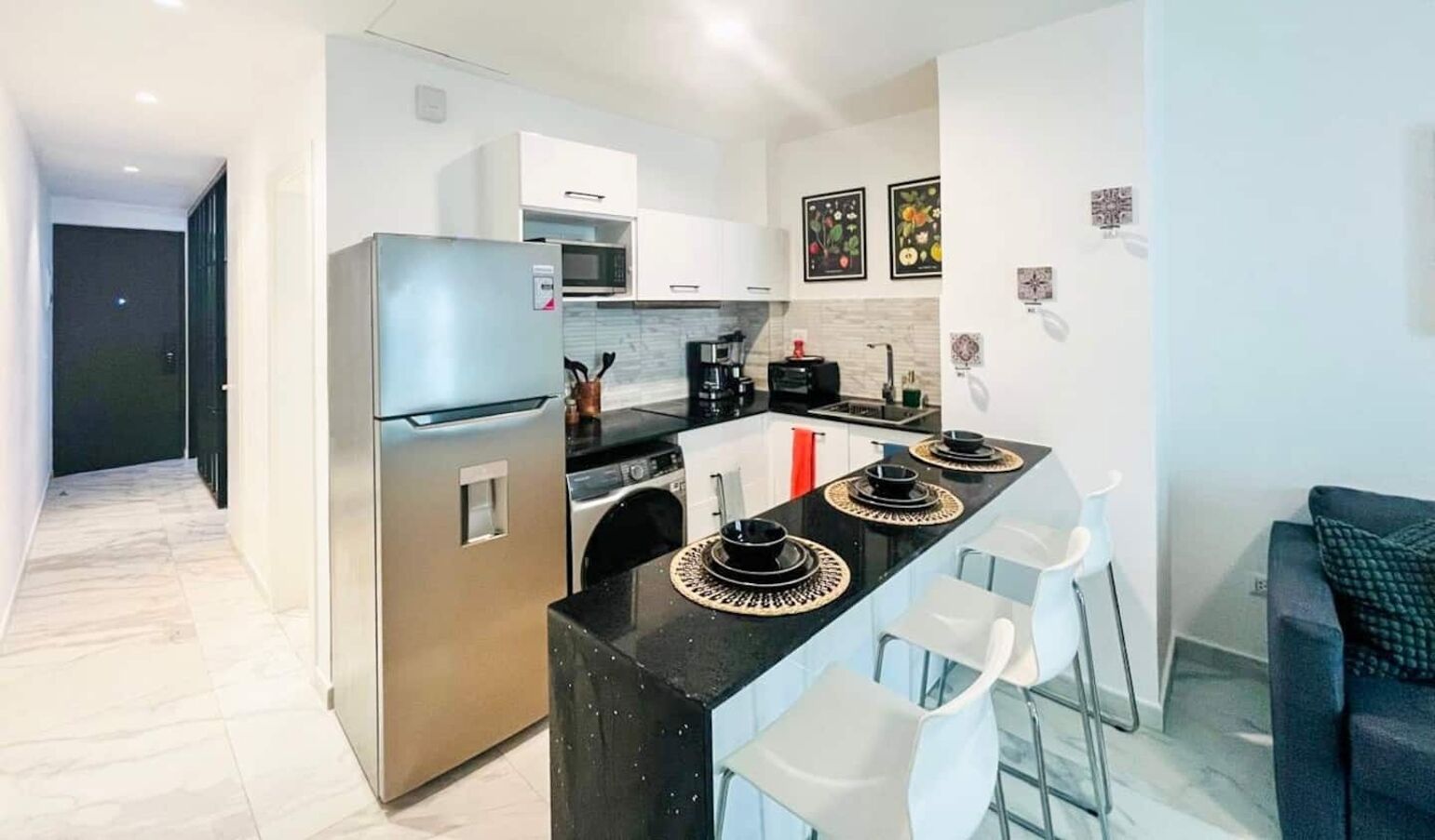 Cocina moderna con refrigerador, microondas y cafetera. Hay sillas blancas y una encimera negra.