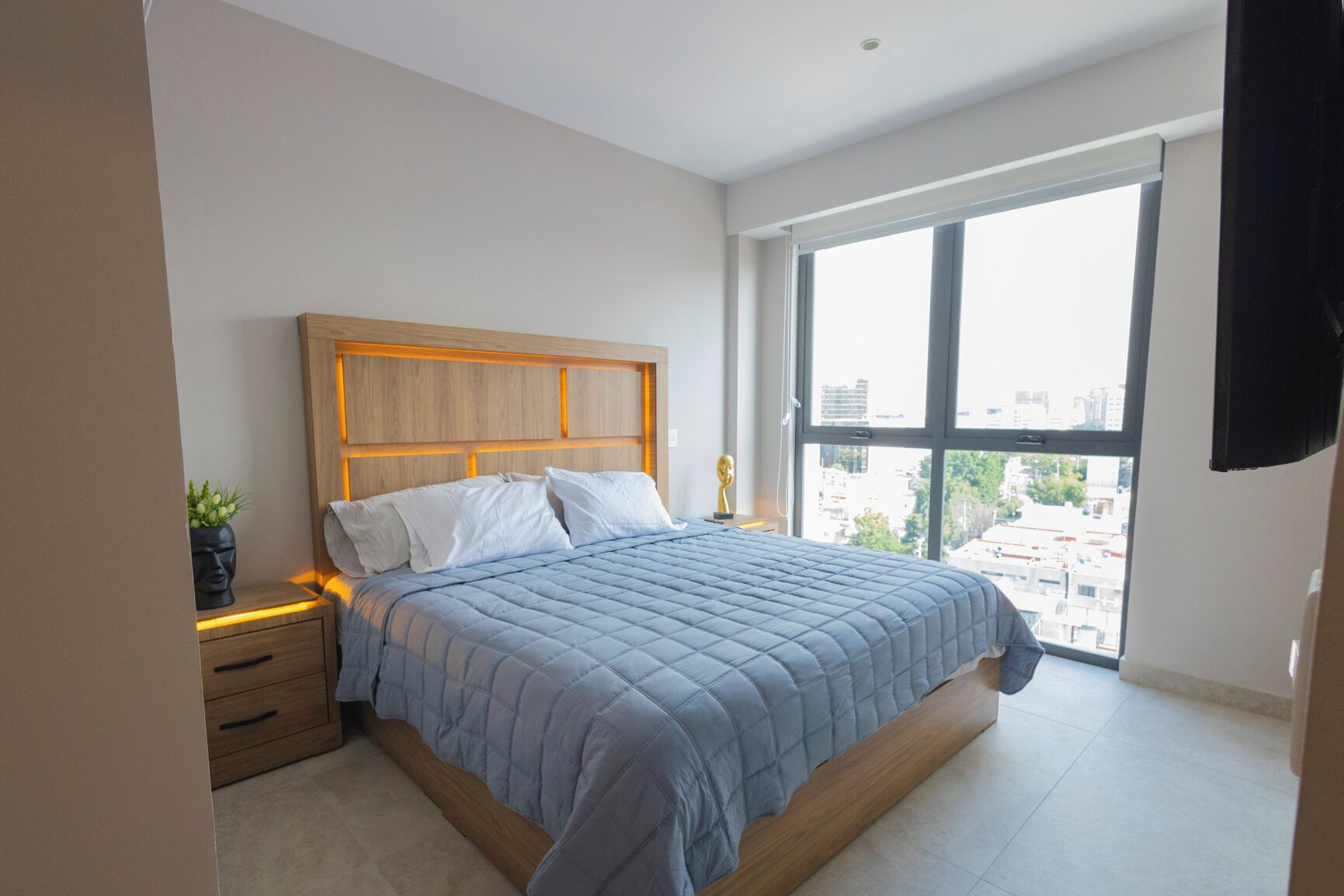 A modern bedroom with a large bed, wooden headboard, bedside tables, and a view of the city through a large window.