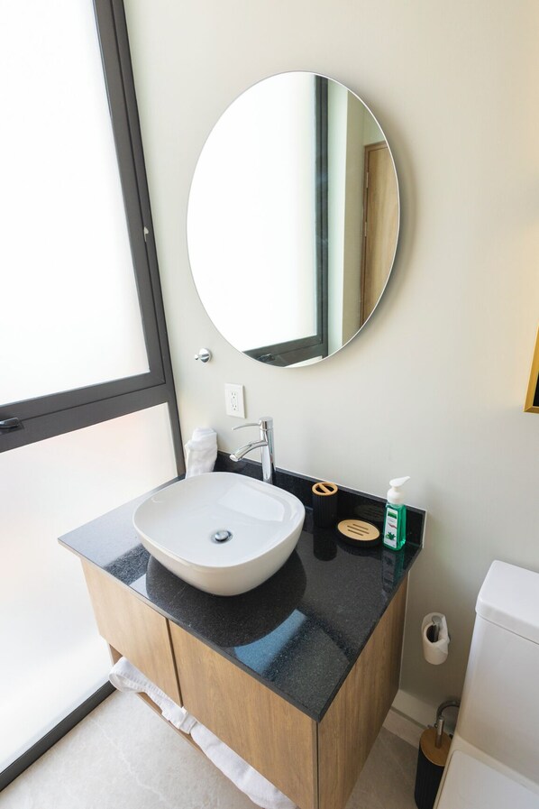 A bathroom with a round mirror, a white sink, a black countertop, and a toilet.