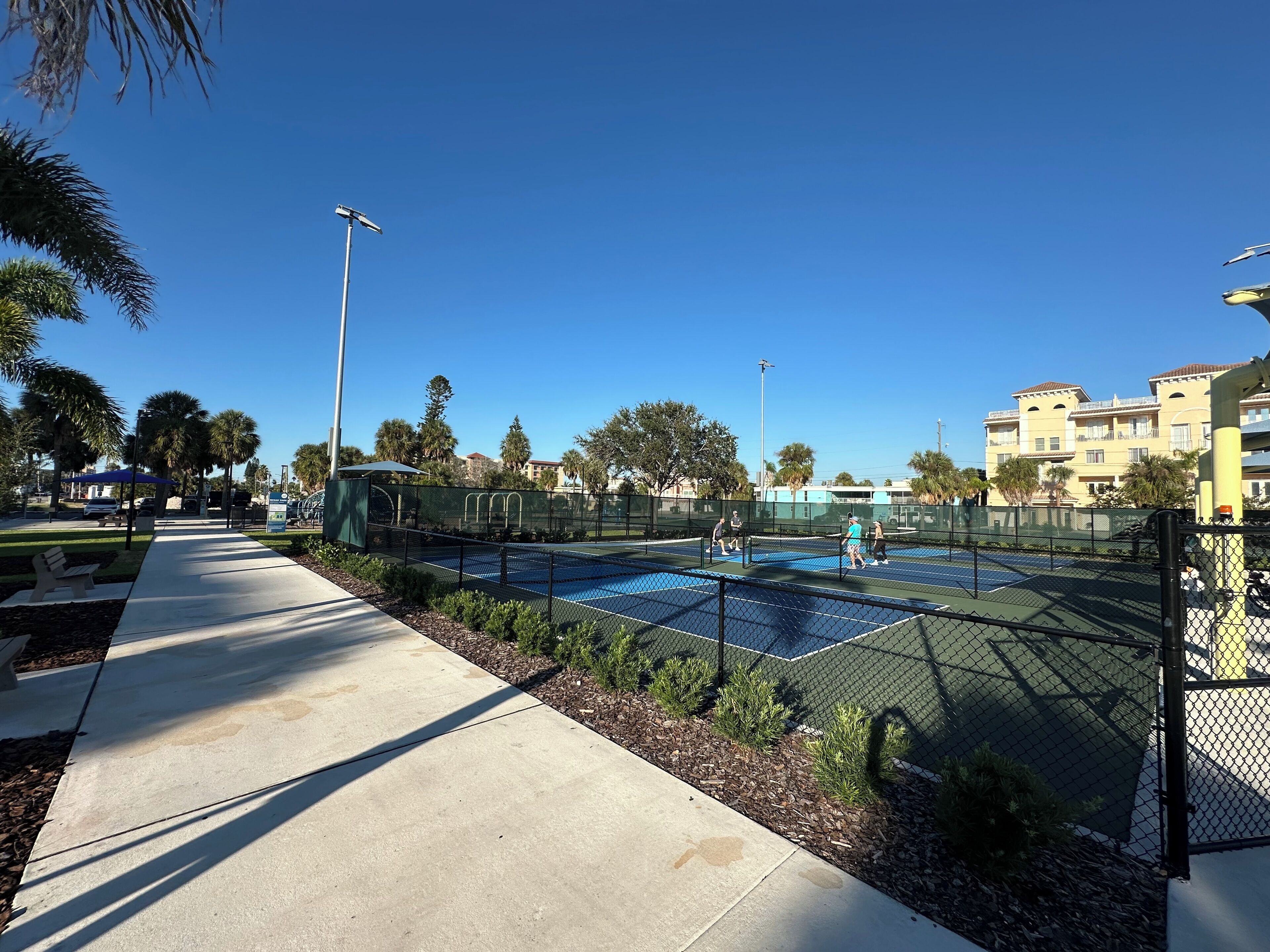 Ein Tennisplatz mit Maschendrahtzaun, umgeben von einem Bürgersteig und kleinen Pflanzen.