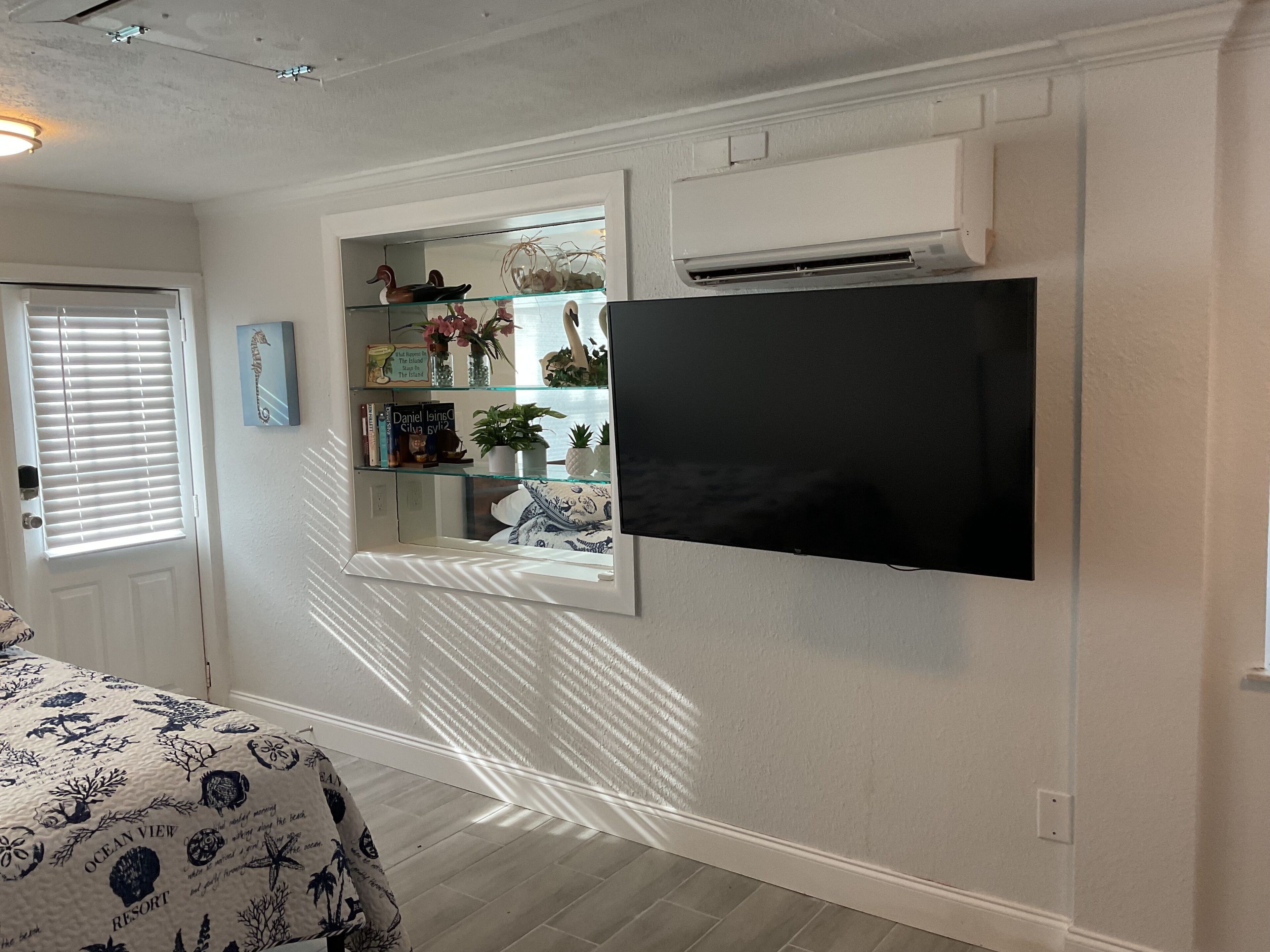 Una habitación con televisión montada, una estantería de vidrio con objetos decorativos, una cama con colcha estampada y un equipo de aire acondicionado.