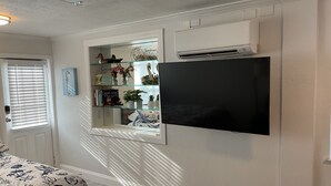 Een kamer met een wandtelevisie, een open kast met decoratieve objecten, een bed met een bedovertrek met dessin, en een airconditioning.