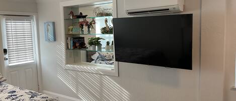 Una habitación con televisión montada, una estantería de vidrio con objetos decorativos, una cama con colcha estampada y un equipo de aire acondicionado.