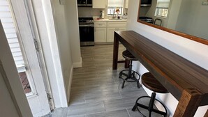 Een smalle gang die leidt naar een keuken met witte kasten, een houten bankje en een grote spiegel.