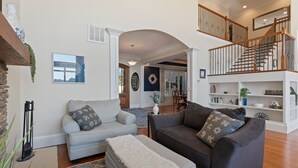 A spacious living room with a grey sofa, a dark sofa, and a white ottoman. There is a staircase with wooden railings leading to an upper level.