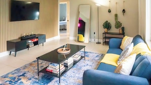 A modern living room with a flat-screen TV, a coffee table with books, a blue sofa with yellow cushions, and a large mirror. - Chic-Miami GoodVibesOnly – Mins to Brickell & Beaches (Miami)