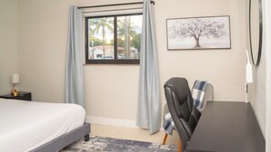 A hotel room with a bed, a desk, a chair, a window with curtains, a wall-mounted picture, and a nightstand with a lamp. - Chic-Miami GoodVibesOnly – Mins to Brickell & Beaches (Miami)