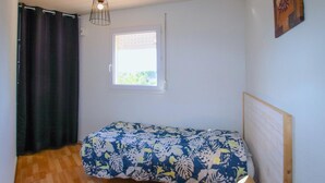 Una camera piccola con un letto singolo, pavimento in legno, una finestra con persiane, un letto con copriletto floreale e un punto luce fissato al muro.