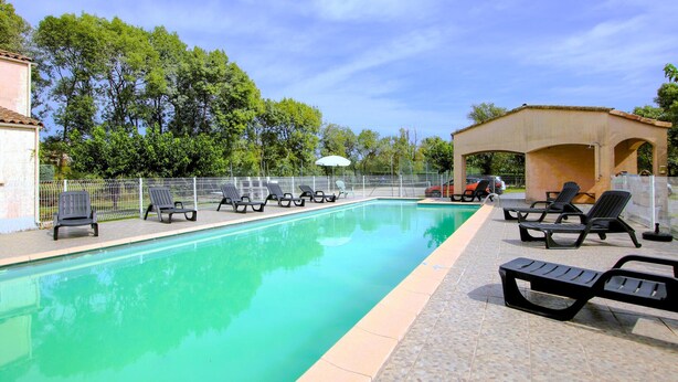 Una piscina con lettini e un'area salotto coperta, circondata da una recinzione e alberi.