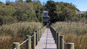 A wooden boardwalk leading to a two-story building with a balcony, surrounded by dense vegetation. - Once Upon a Tide: Stunning Soundfront, Private Pool, Hot Tub, Private Dock (Corolla)