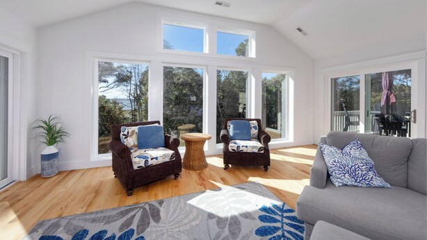 A spacious living room with high ceilings and large windows, featuring wicker chairs, a grey sofa, and a wooden floor. - Once Upon a Tide: Stunning Soundfront, Private Pool, Hot Tub, Private Dock (Corolla)