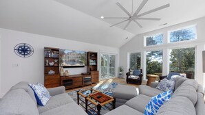 A spacious living room with a large gray sectional sofa, a wooden coffee table, and a ceiling fan. There is a bookshelf with books and decorative items, and a large window offering a view of trees and a building. - Once Upon a Tide: Stunning Soundfront, Private Pool, Hot Tub, Private Dock (Corolla)