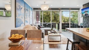 A modern living room with a wooden dining table, a sofa, and a large window overlooking a patio area. - 2BR Walk to Ski - Spa-like Bath (Park City)