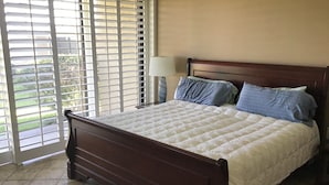 A bedroom with a wooden bed, a nightstand with a lamp, and a view through a window with blinds. - 30 days+, 2-bedroom condo in Palm Desert, California, with view, hot tub & pool (Palm Desert)