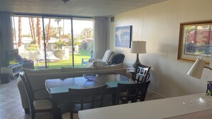 A living room with a dining table, chairs, a sofa, a TV, and a large window with a view of a garden. - 30 days+, 2-bedroom condo in Palm Desert, California, with view, hot tub & pool (Palm Desert)
