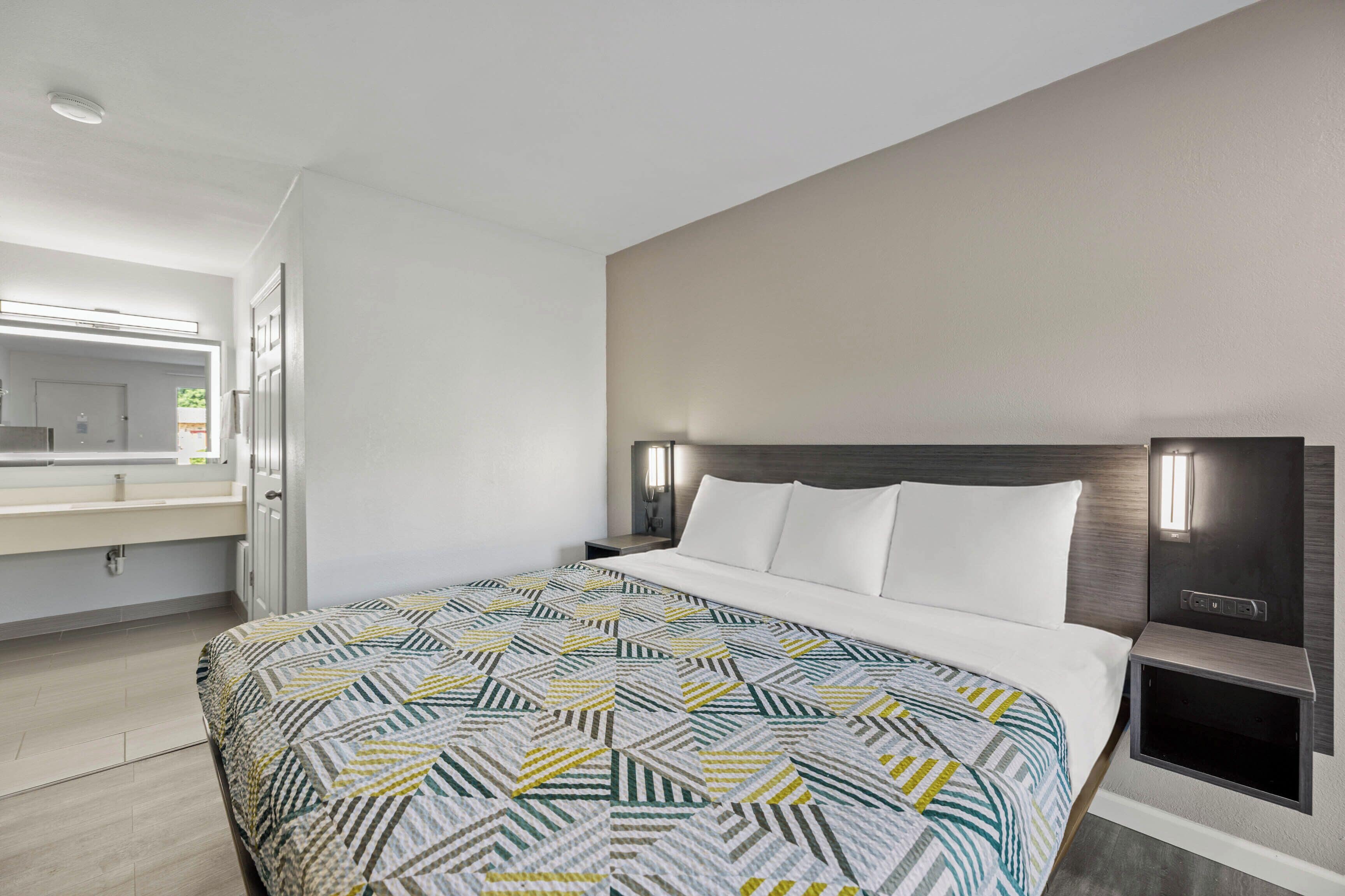 A hotel room with a bed, a nightstand, a bedside table, a mirror, and a window.