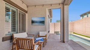 A covered patio with wooden furniture, a pool, and a painting of a lake. - Verrado Views Retreat - Sleeps 12 + Pool (Buckeye)