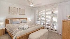 Een slaapkamer met een houten bed, een bankje, een nachtkastje, een plafondventilator en een raam met jaloezieën.