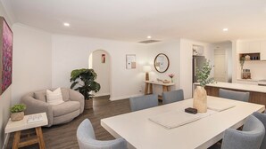 Een moderne woonkamer met een bank, een eettafel met stoelen, een grote plant en een keuken in de achtergrond.