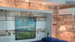 A bedroom with a white bed, a blue bedspread, a painting of a landscape, and a lamp on the wall. - Tiny house 5 minutes near Kemah boardwalk! (Bacliff)
