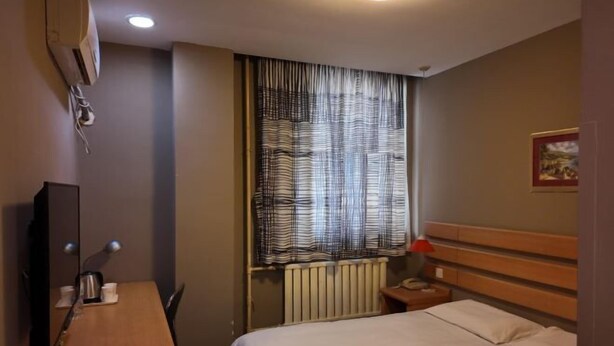 A hotel room with a bed, a desk, a chair, a television, a window with curtains, a radiator, and a nightstand.
