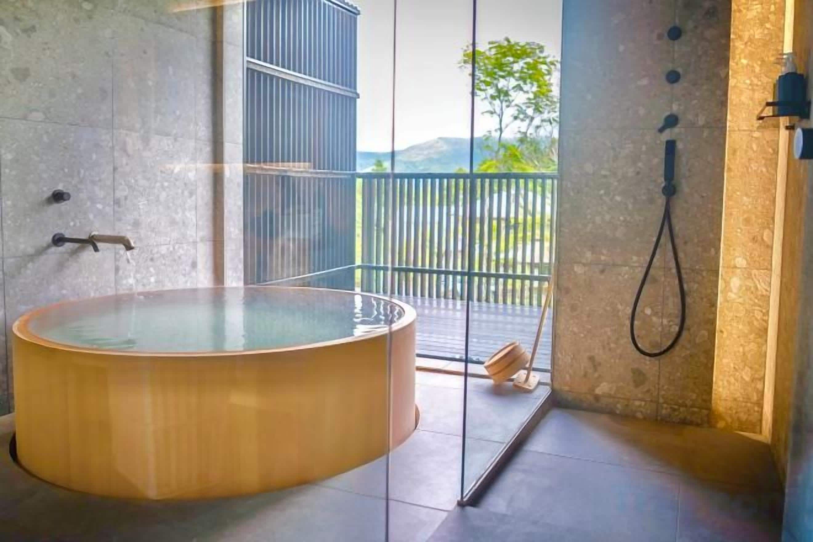 A modern bathroom with a large wooden bathtub, a glass shower enclosure, and a view of the outdoors.