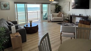A living room with a large TV, a ceiling fan, a sofa, a coffee table, and a view of the ocean through the sliding doors. - The Brunswick Penthouse with Panoramic Oceanfront Balconies-2 bedroom/2.5 bath (Old Orchard Beach)